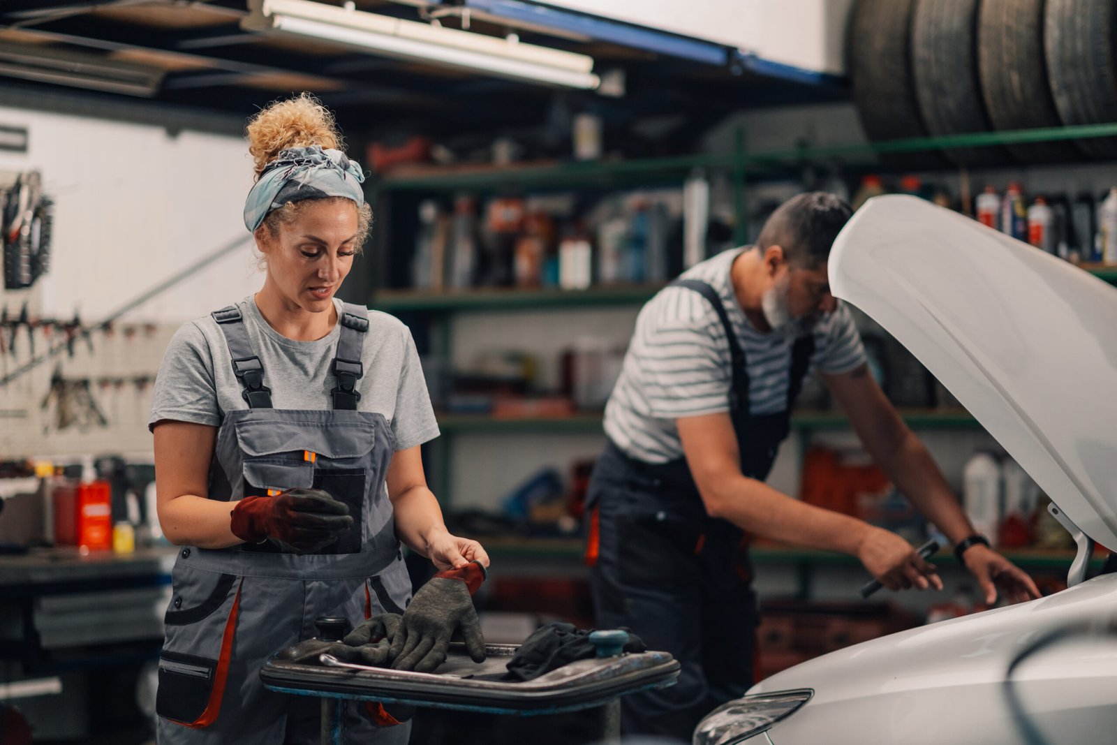 Professional female auto mechanic in uniform is standing at car repair shop and putting working gloves on while repairman in a blurry background is looking under car hood and fixing it. Copy space.
